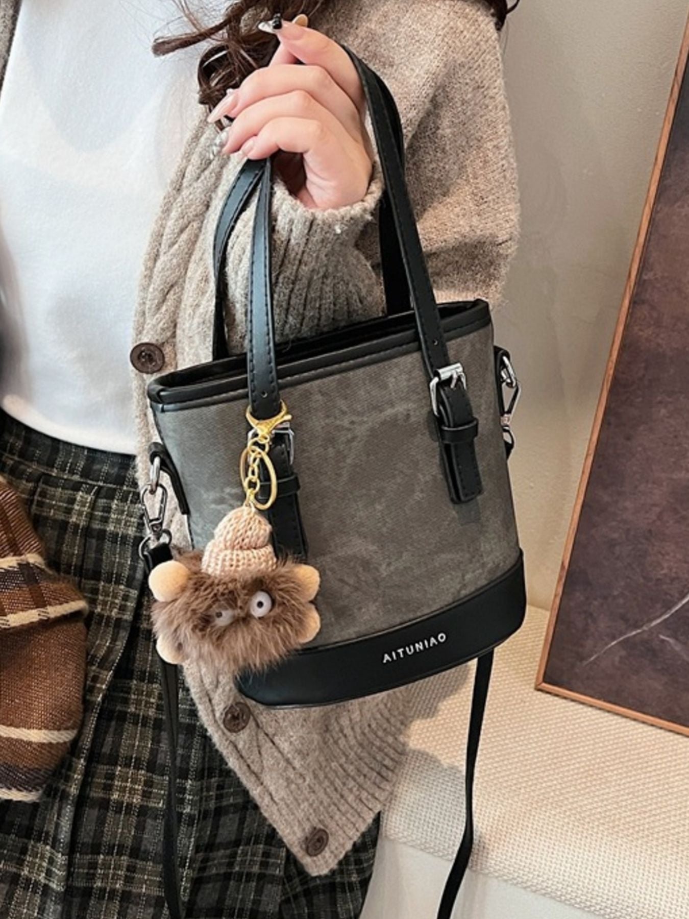 Person holding a gray handbag with a fluffy keychain, wearing a plaid skirt and beige sweater.