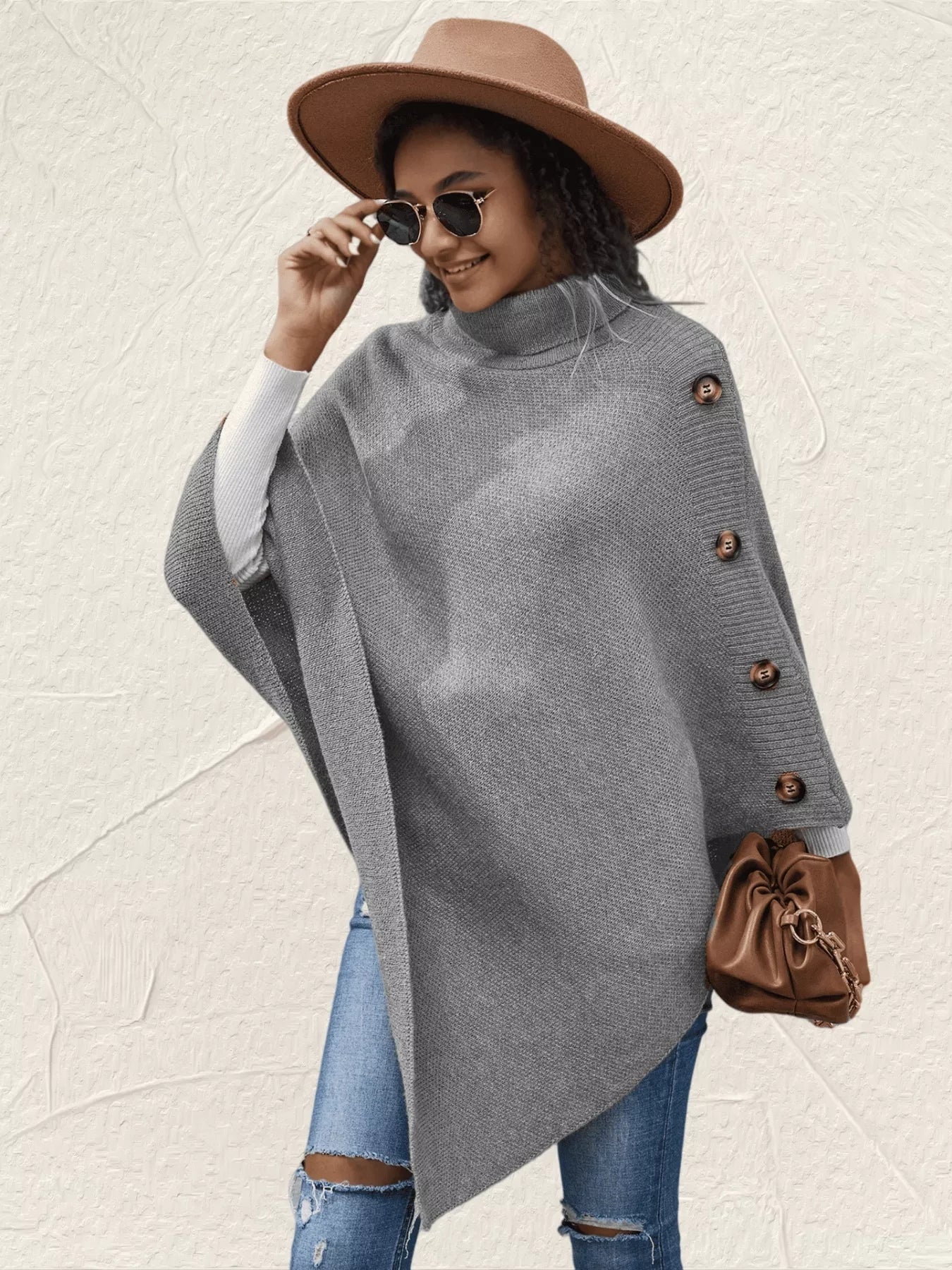 Woman wearing a gray poncho with brown buttons, blue jeans, and a brown hat on a light background