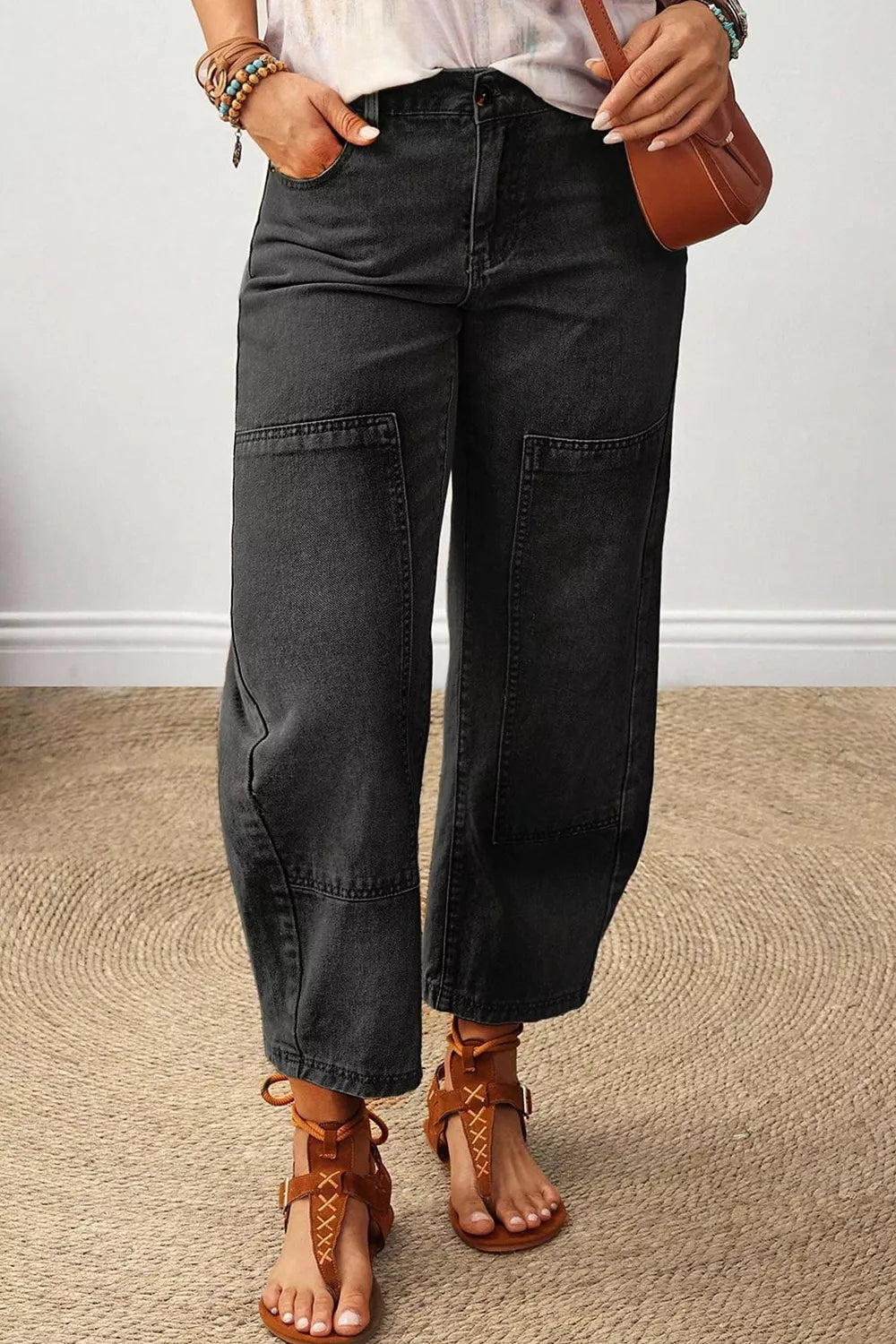 A person wearing dark charcoal gray wide-leg patchwork jeans, styled with brown lace-up sandals, bracelets, and a small brown shoulder bag, standing on a textured beige rug.