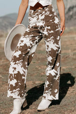 A person outdoors wearing high-waisted wide-leg pants with a brown and off-white cow-print pattern, holding a light-colored hat and wearing white cowboy boots.