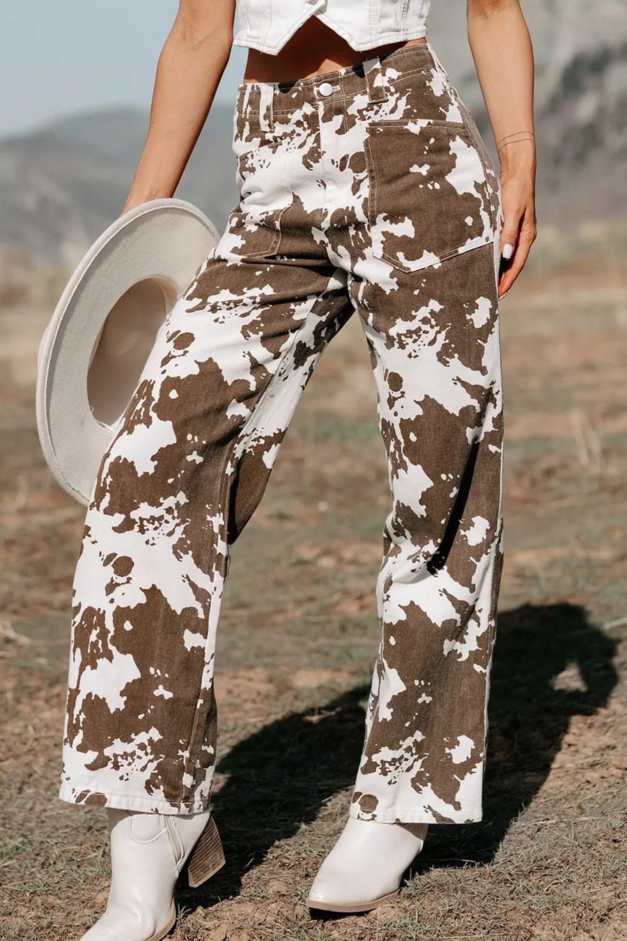A person outdoors wearing high-waisted wide-leg pants with a brown and off-white cow-print pattern, holding a light-colored hat and wearing white cowboy boots.