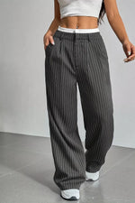 A person wearing loose, wide-leg pinstripe pants in dark gray with thin white vertical stripes, paired with a white crop top and white sneakers, standing against a light gray wall.