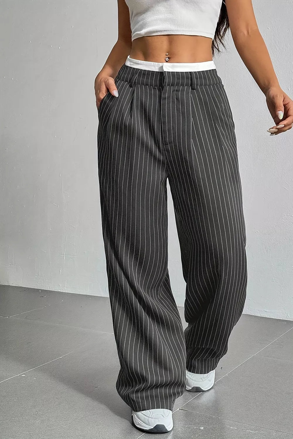 A person wearing loose, wide-leg pinstripe pants in dark gray with thin white vertical stripes, paired with a white crop top and white sneakers, standing against a light gray wall.
