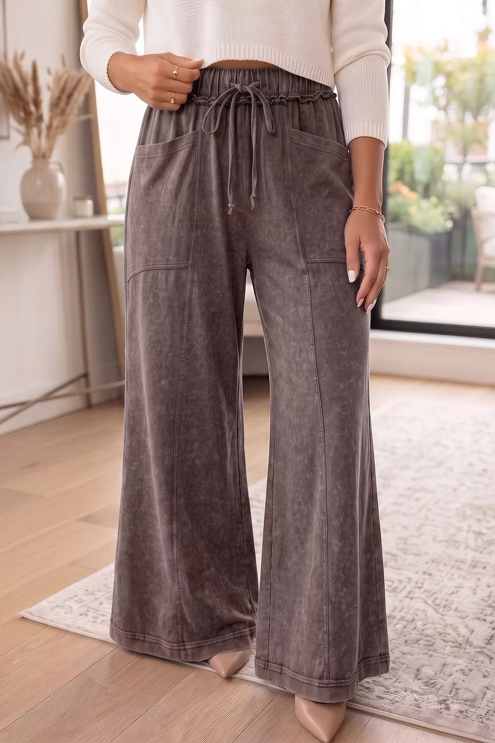 A person wearing wide-leg drawstring pants in a dark gray washed fabric, paired with a cream sweater and nude heels, standing in a bright indoor room with wood floors and a rug.
