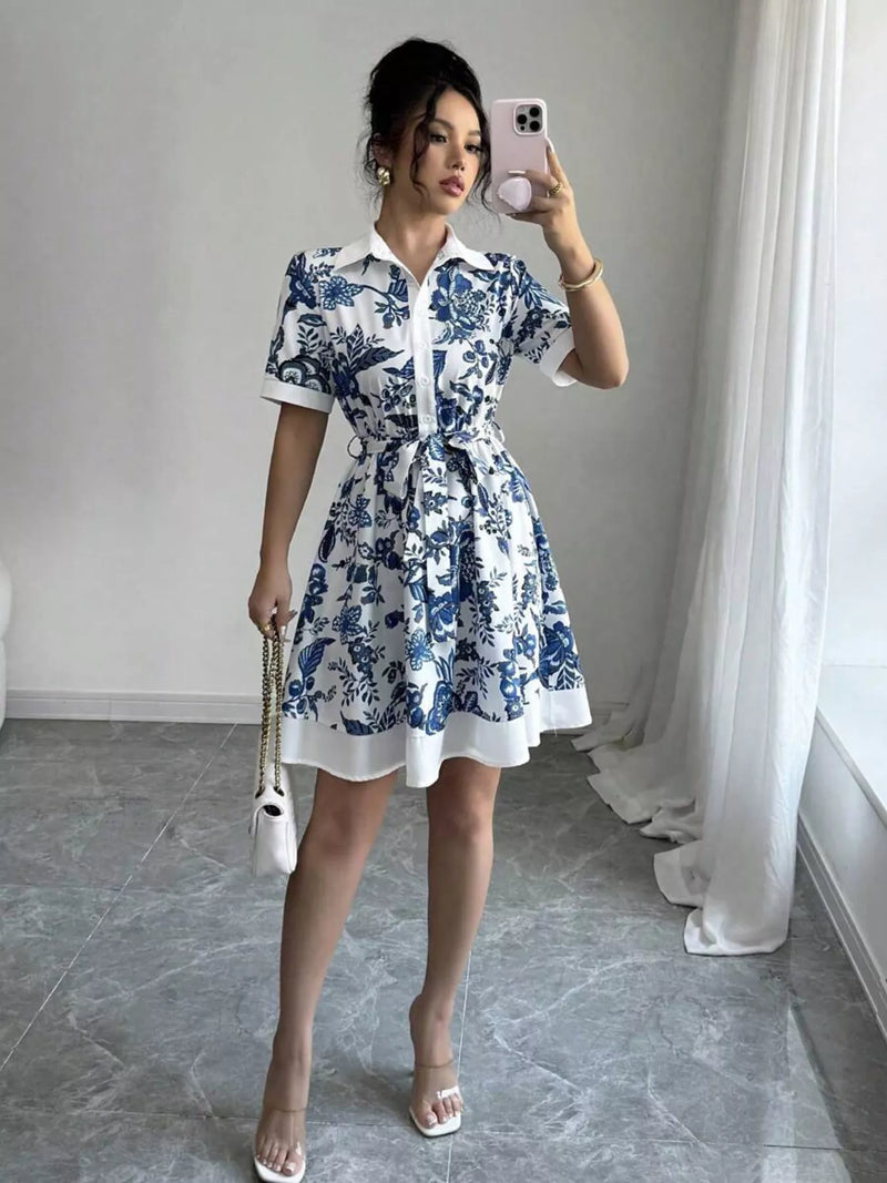 Woman taking a mirror selfie wearing a blue and white floral dress in a room with gray flooring and white curtains.