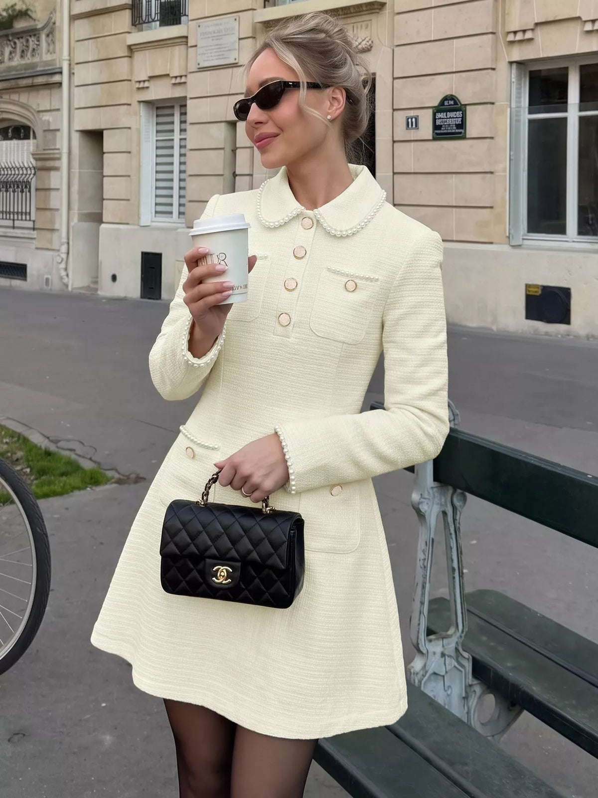 A woman wearing a cream-colored tweed mini dress with pearl collar and gold buttons, holding a coffee cup and a black quilted handbag with a gold logo, standing on a city street next to a bench.