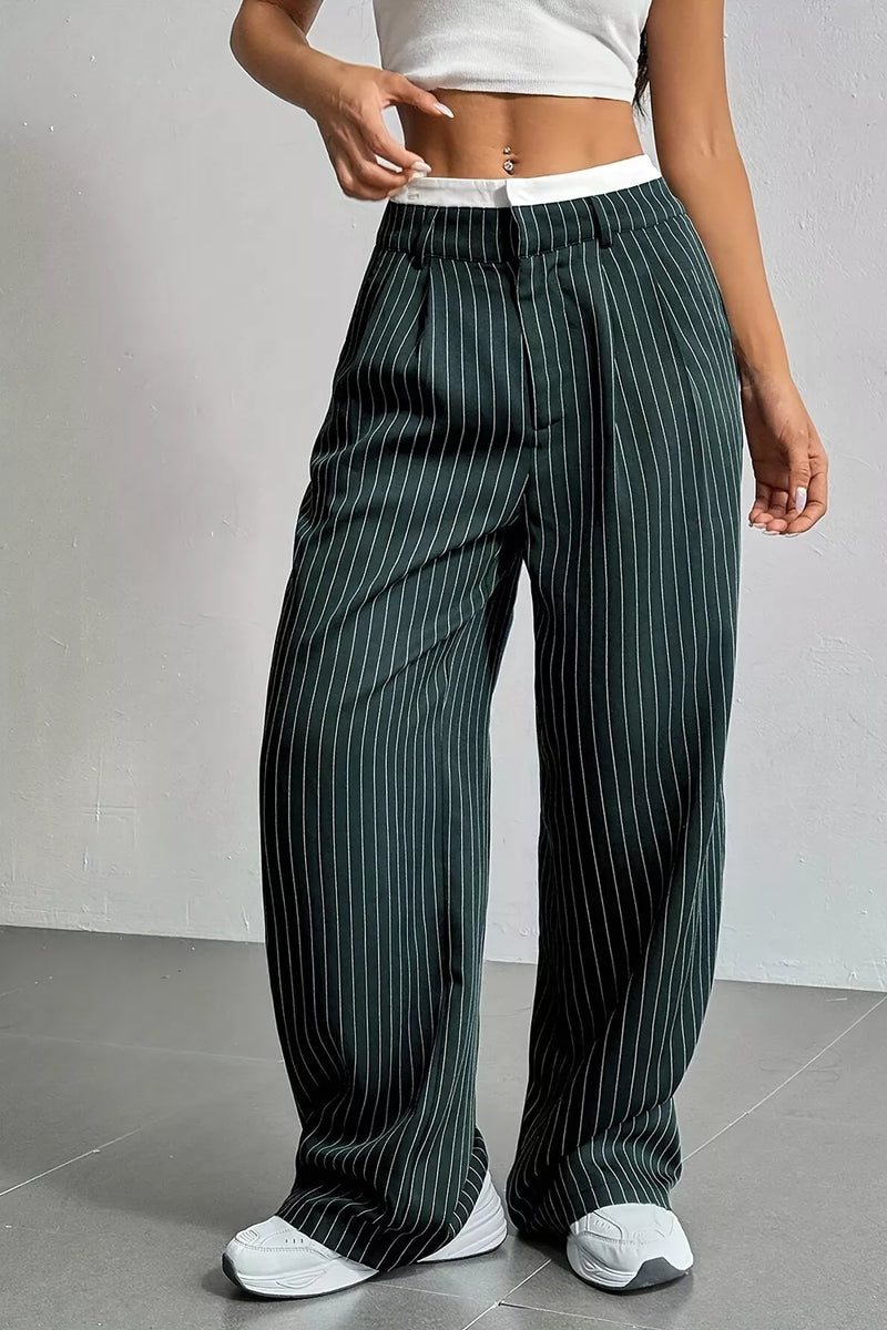 A person wearing dark green wide-leg pinstripe trousers with a white waistband, paired with a white crop top and white sneakers, standing against a light gray wall.