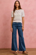Woman wearing a white lace top and blue jeans against a pink background