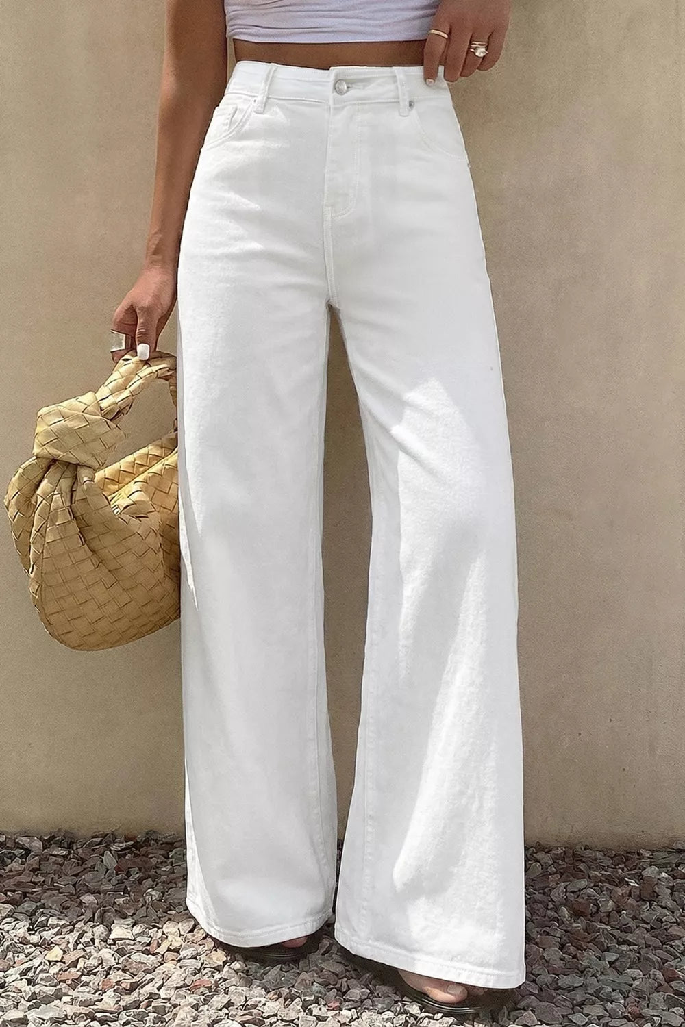 Person wearing white wide-leg jeans holding a straw bag against a beige wall.