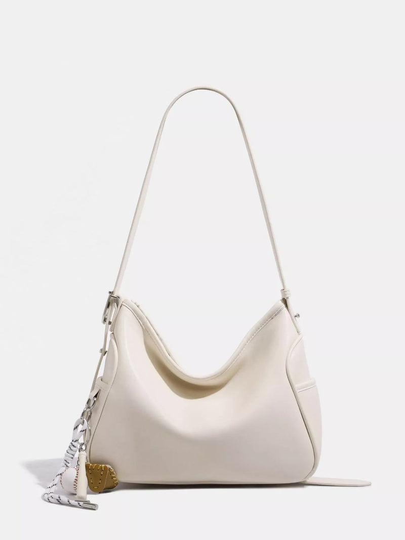An off-white leather slouchy shoulder bag with a thin matching strap, silver-tone hardware, and a decorative silver chain tassel with a gold heart charm, shown on a light gray background.