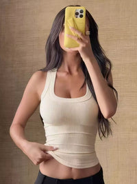 Woman taking a mirror selfie wearing a beige tank top against a beige wall.
