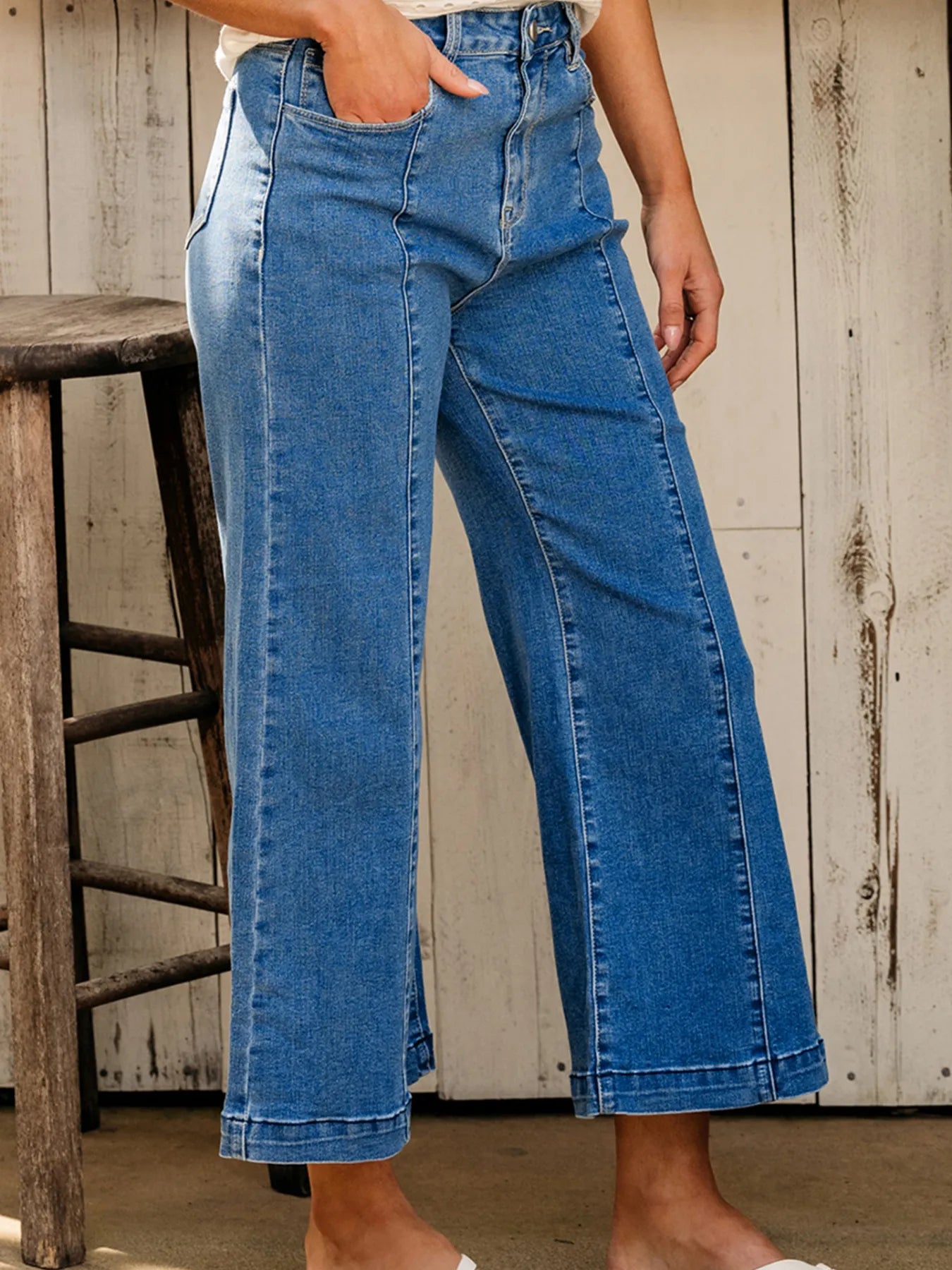 Person wearing blue jeans standing next to a wooden stool against a rustic wall.
