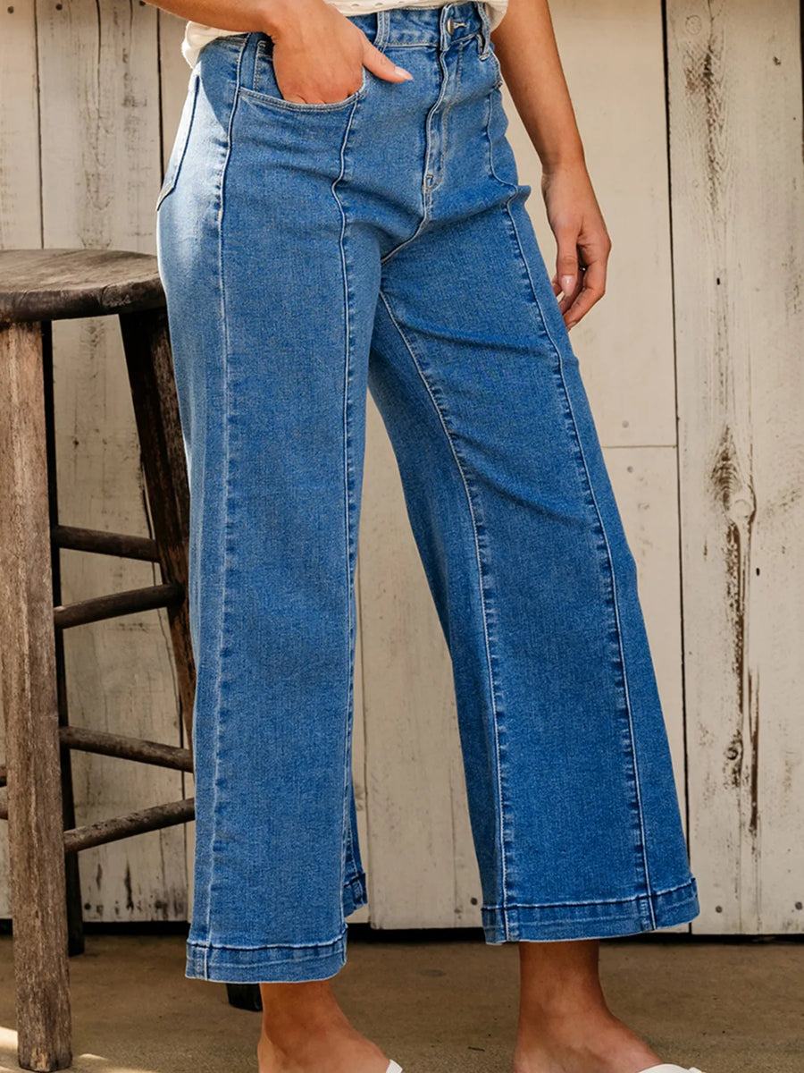 Person wearing blue jeans standing next to a wooden stool against a rustic wall.