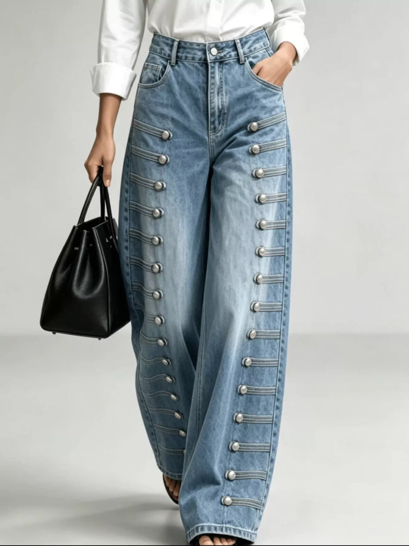 A person wearing light blue wide-leg denim jeans with decorative silver-tone military-style button and cord details down the front, paired with a white top and holding a black handbag against a light gray studio background.