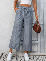 Person wearing striped wide-leg pants and white sneakers, holding a brown handbag against a wooden panel background.