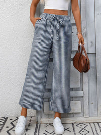 Person wearing striped wide-leg pants and white sneakers, holding a brown handbag against a wooden panel background.