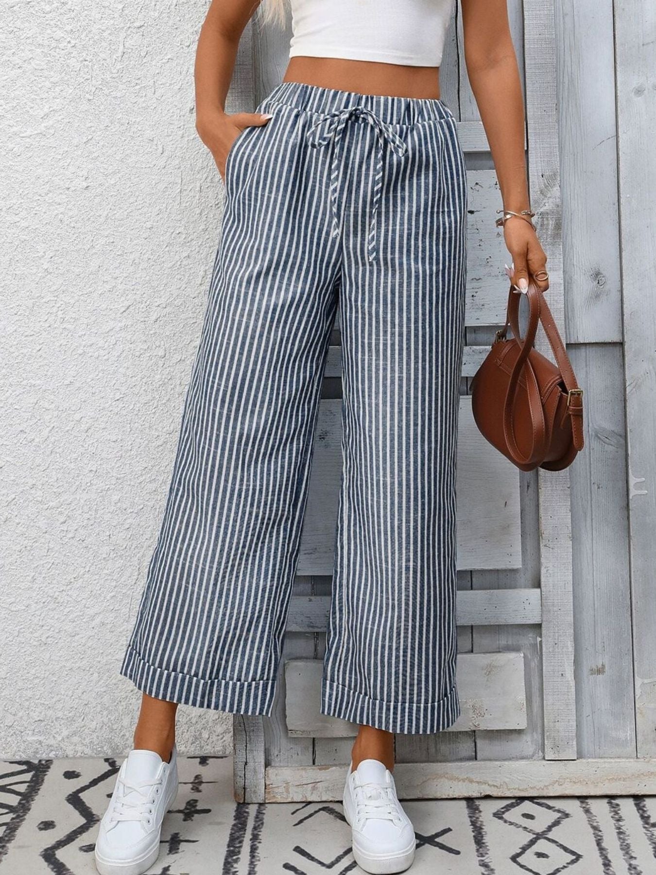 Person wearing striped wide-leg pants and white sneakers, holding a brown handbag against a wooden panel background.