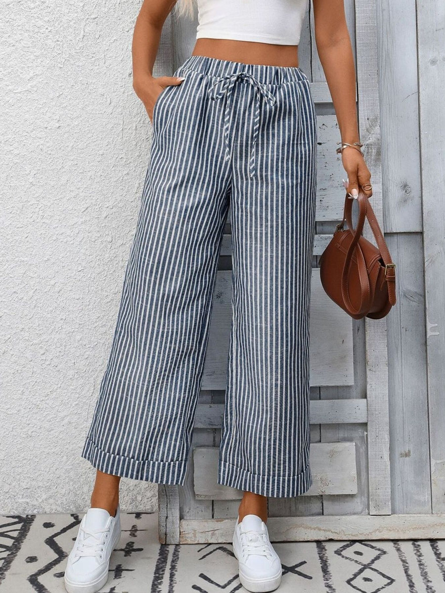 Person wearing striped wide-leg pants and white sneakers, holding a brown handbag against a wooden panel background.