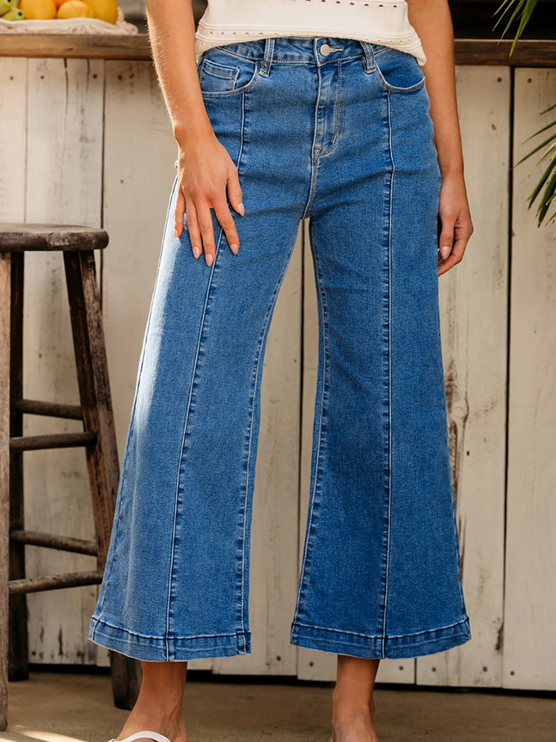 Person wearing blue jeans standing in a rustic setting with wooden furniture and a white wall.