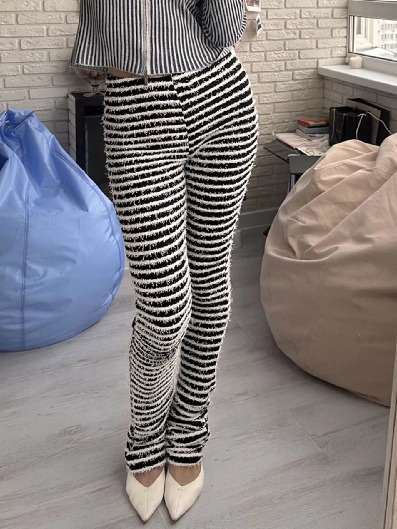 A person wearing black and white horizontally striped textured knit pants, paired with a striped long-sleeve top and white pointed-toe heels, standing in a room with bean bags and a light brick wall.