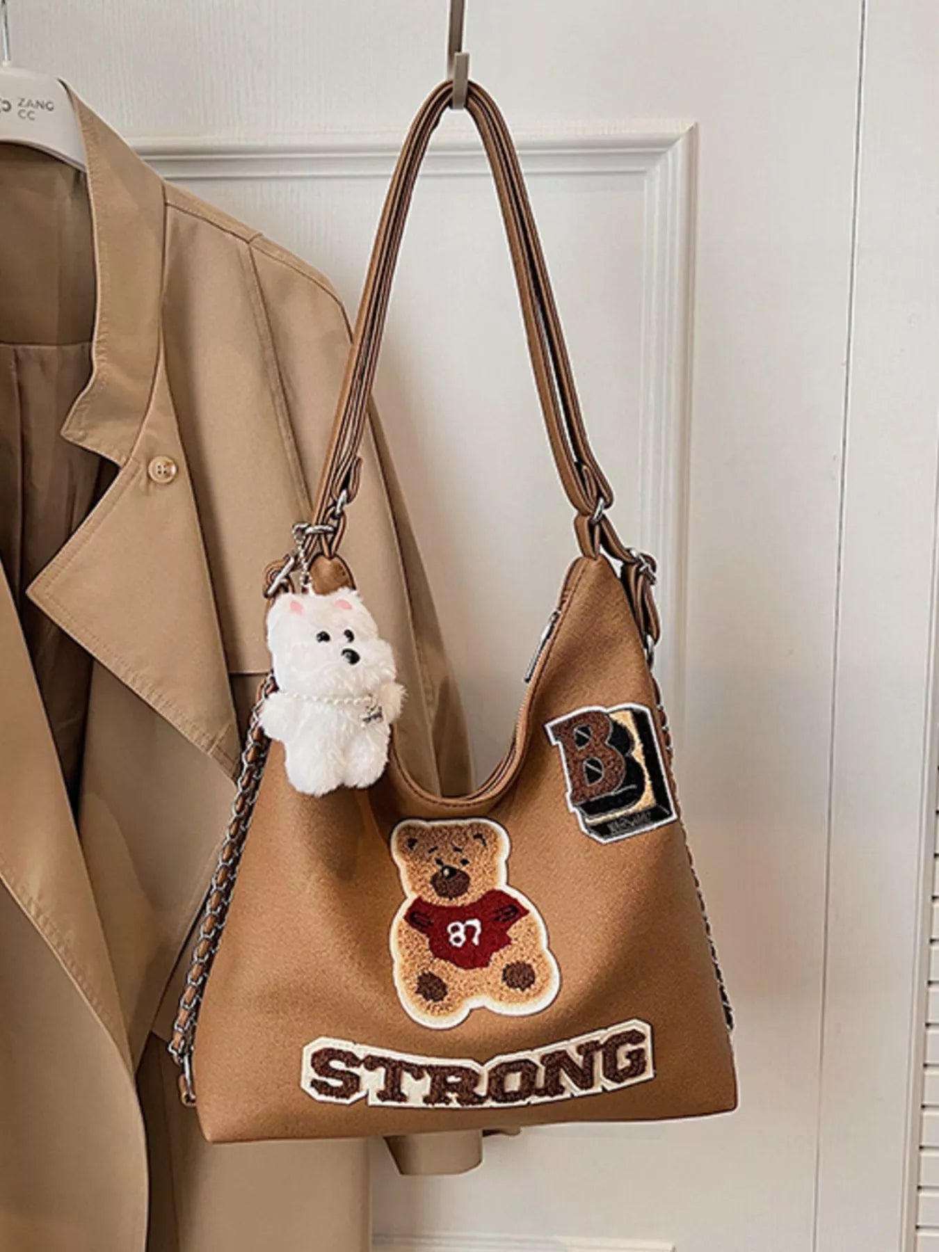 Brown handbag with teddy bear and 'B Strong' patch, hanging on a hook.