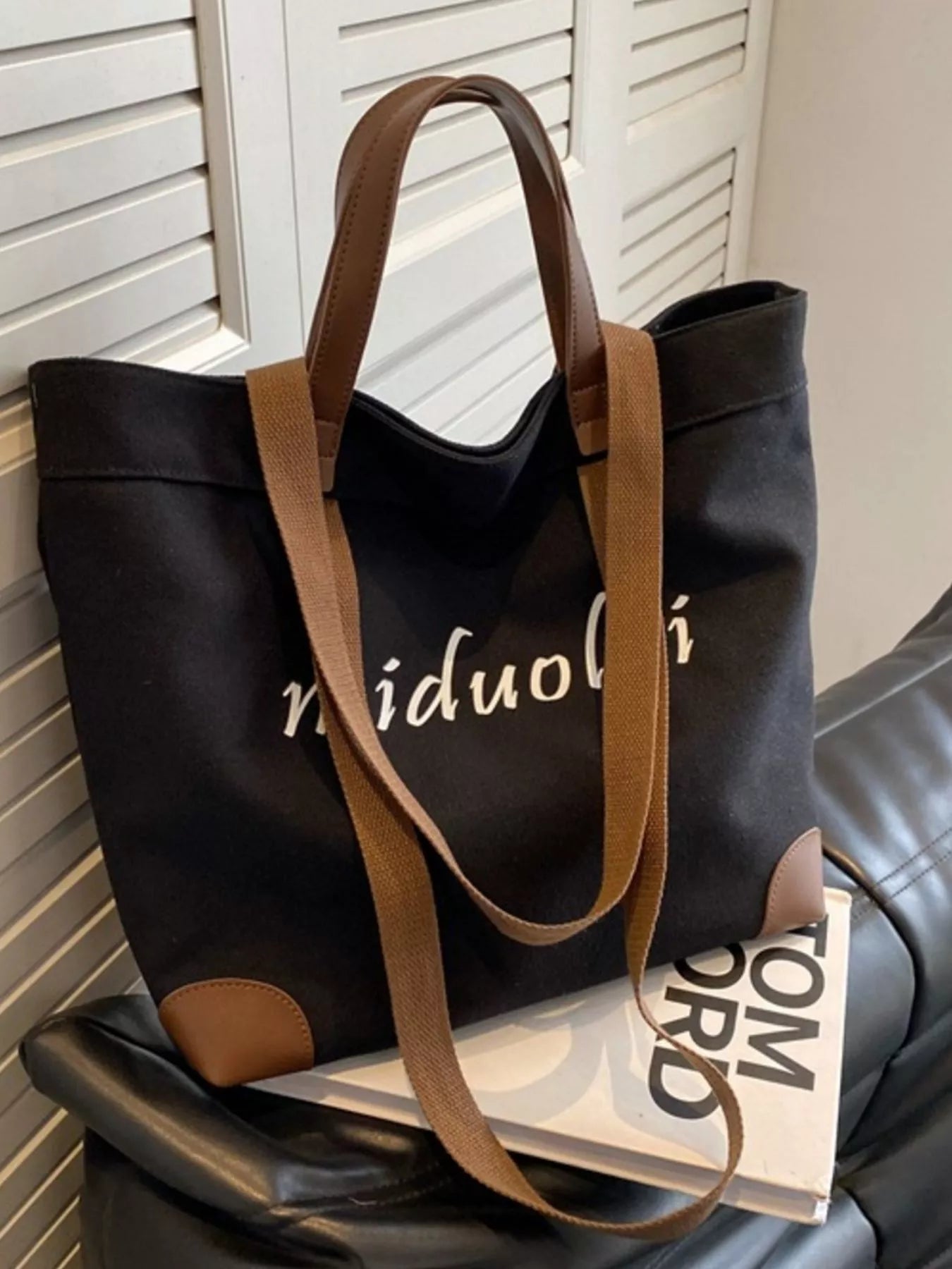 Black tote bag with brown handles on a chair next to a book