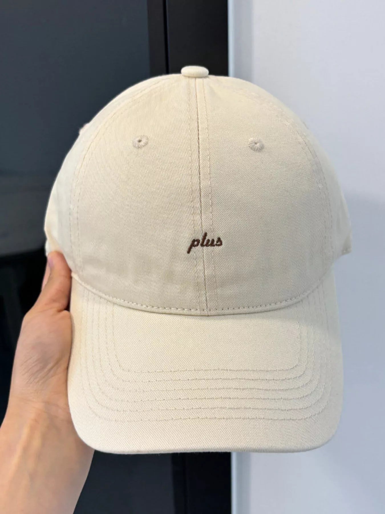 Beige cap with 'plus' embroidery held by a hand against a neutral background