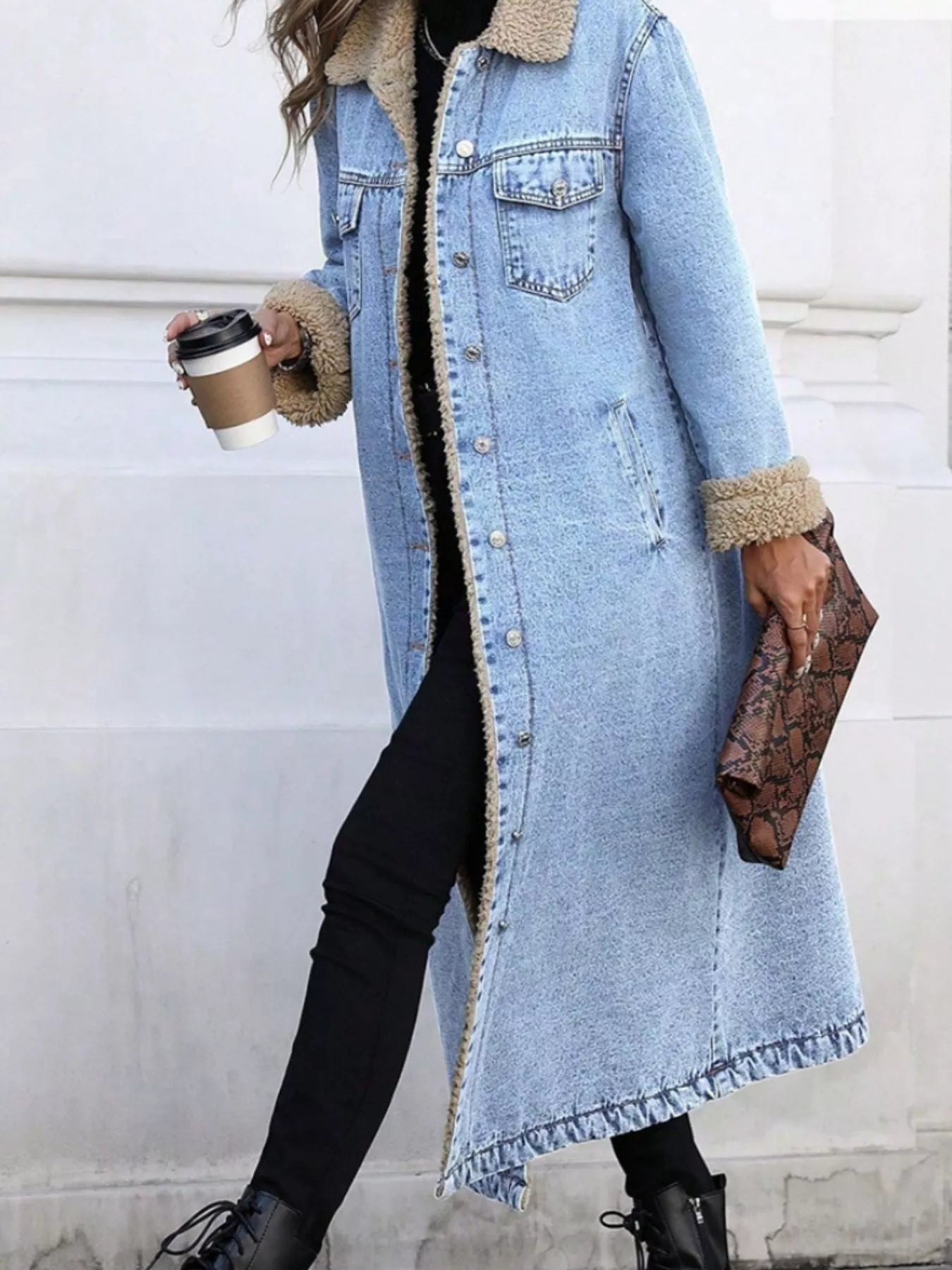 Person wearing a long denim coat with fur trim, holding a coffee cup against a light-colored wall.