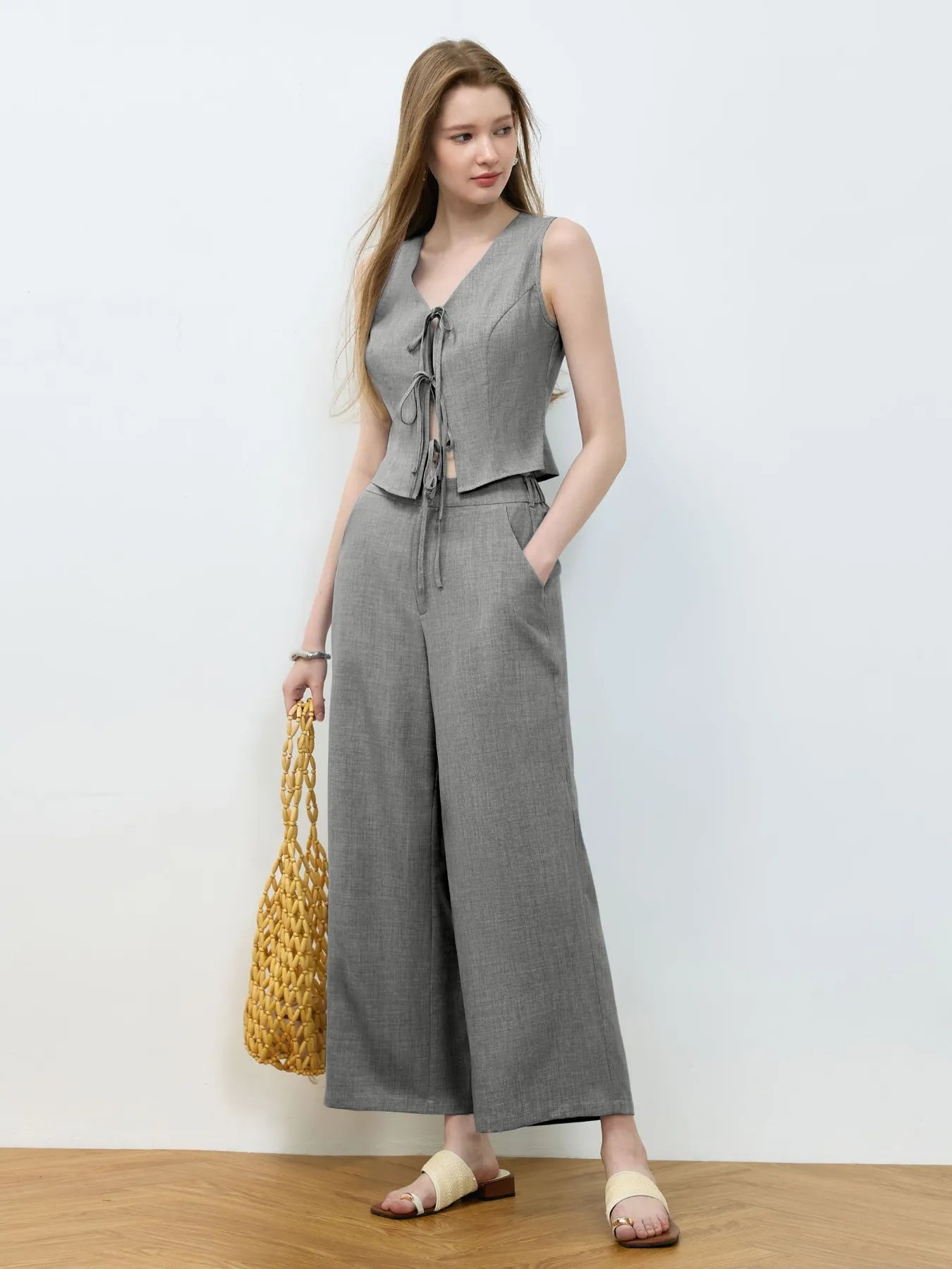 Woman wearing a gray sleeveless top and wide-leg pants, holding a yellow bag against a white background.