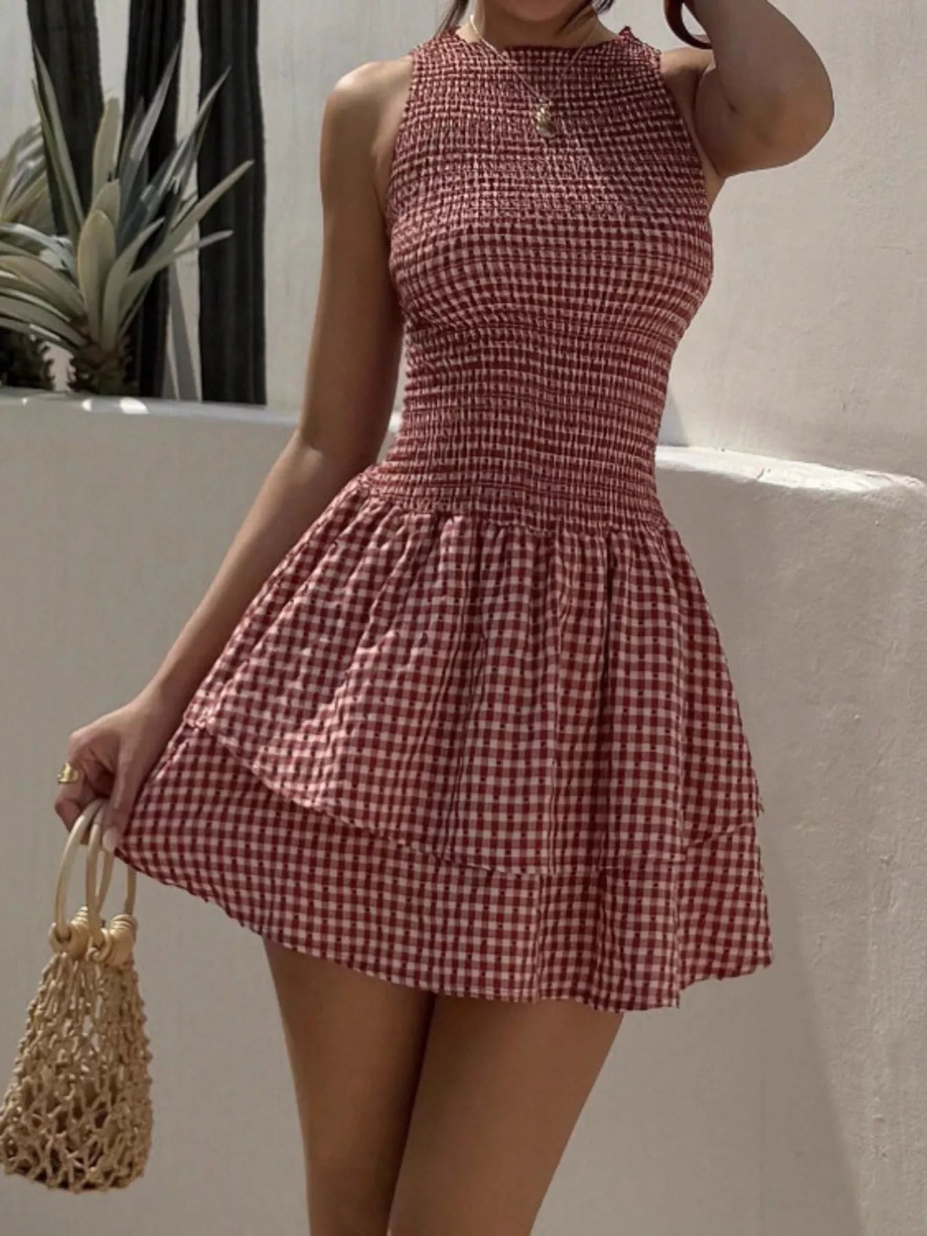 Woman wearing a red and white checkered dress holding a straw bag against a neutral background