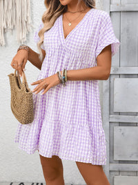 Woman wearing a purple checkered dress holding a woven handbag against a wooden wall.