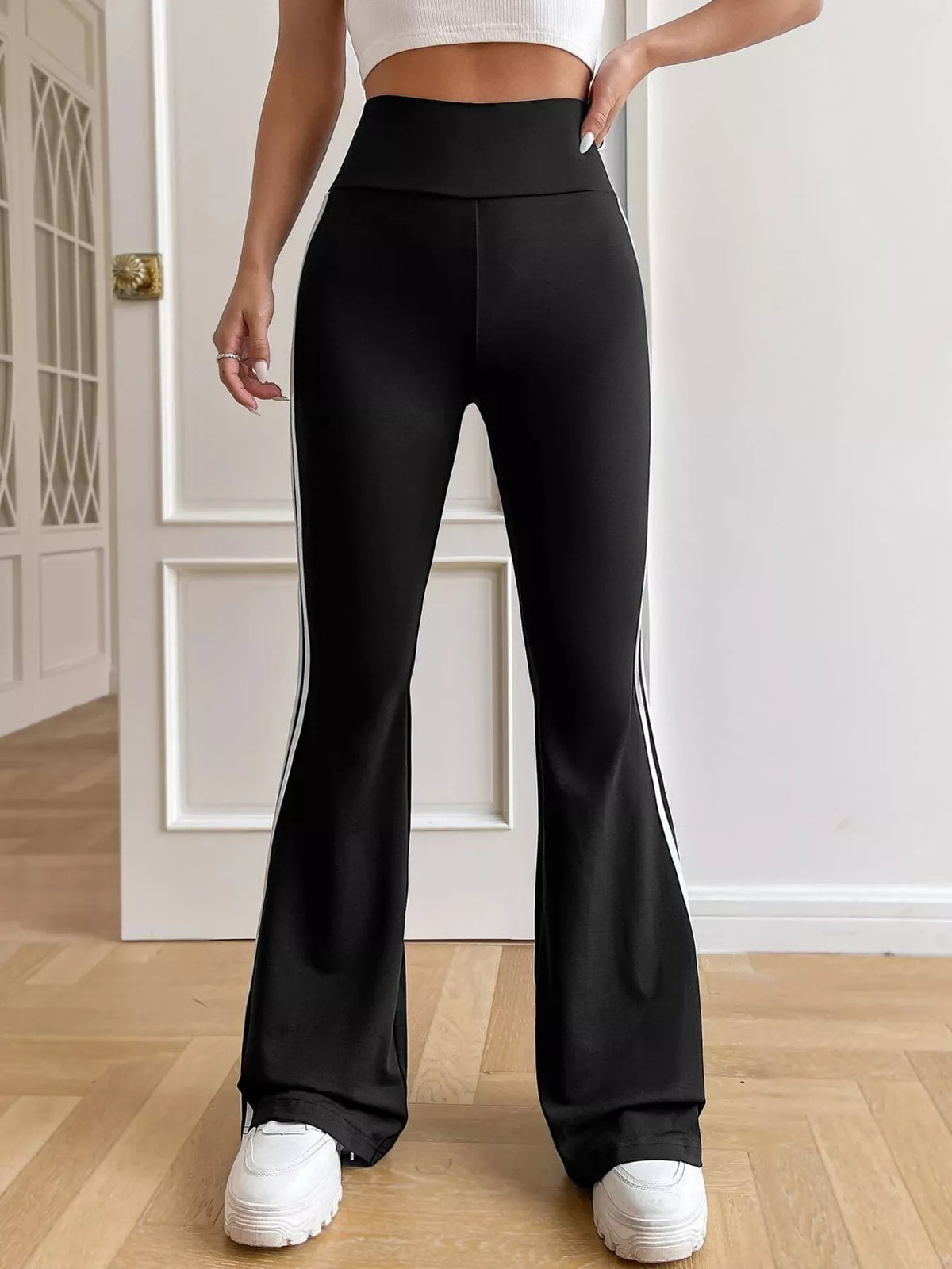 Person wearing black flared pants and white sneakers in a room with wooden floor and white door.