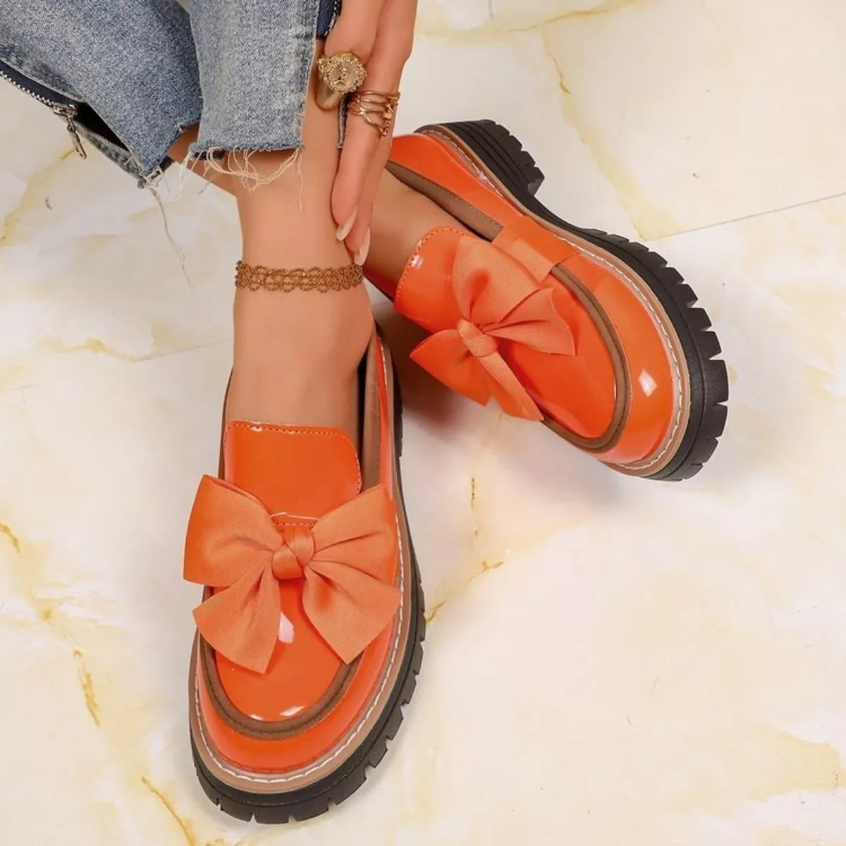 A pair of bright orange patent leather loafers with large satin bows and chunky black lug soles, shown being worn on a person's feet on a light marble floor.