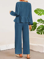 Person wearing a blue peplum top and wide-leg pants with a white wall and green leaves in the background.