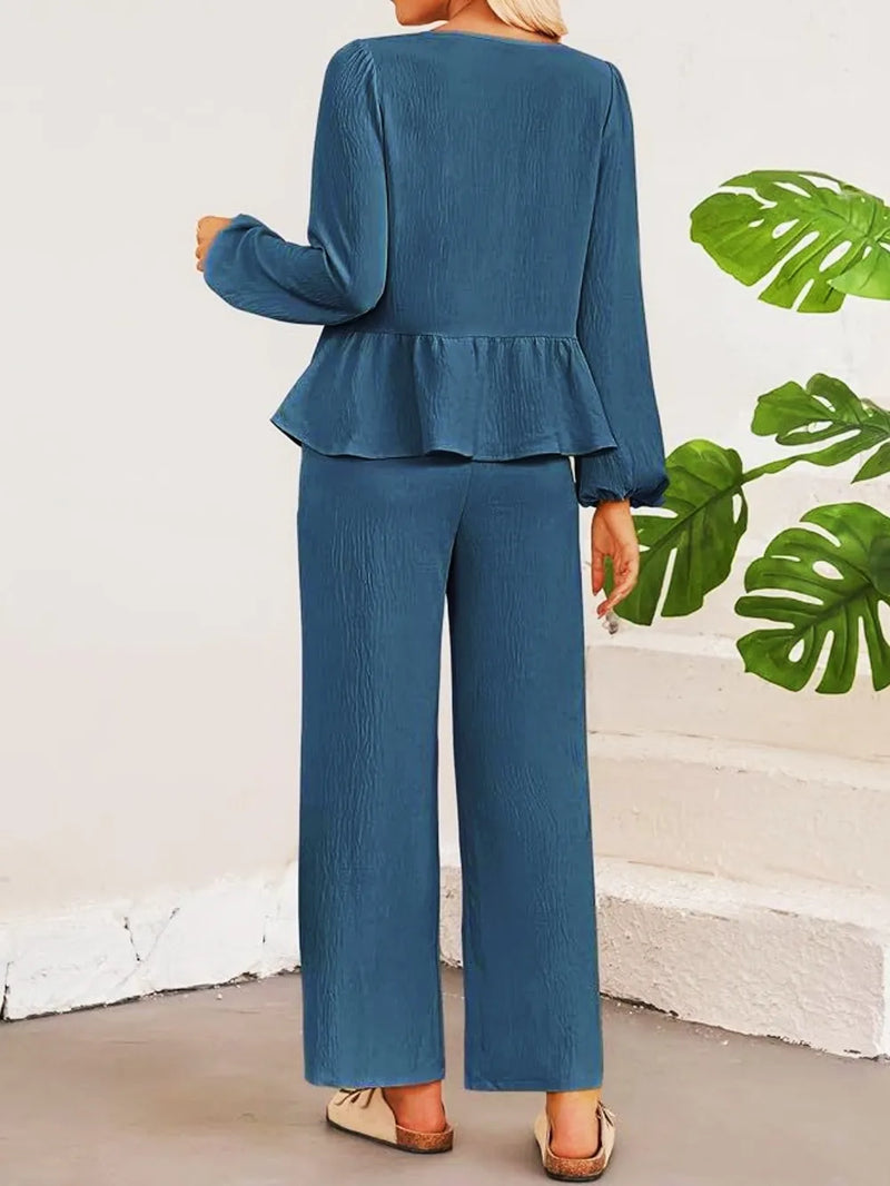 Person wearing a blue peplum top and wide-leg pants with a white wall and green leaves in the background.