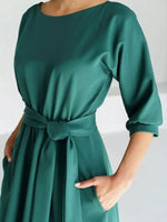 A woman wearing a satin-like dark green dress with elbow-length sleeves, a round neckline, and a matching waist tie, shown against a plain light background.