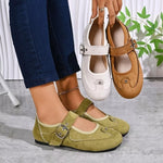 A person wearing olive green Mary Jane-style flats is shown from the knees down, holding up two additional pairs of the same shoe style in white and tan. The shoes have a rounded toe, a decorative cutout detail on the instep, and a silver buckle strap closure.