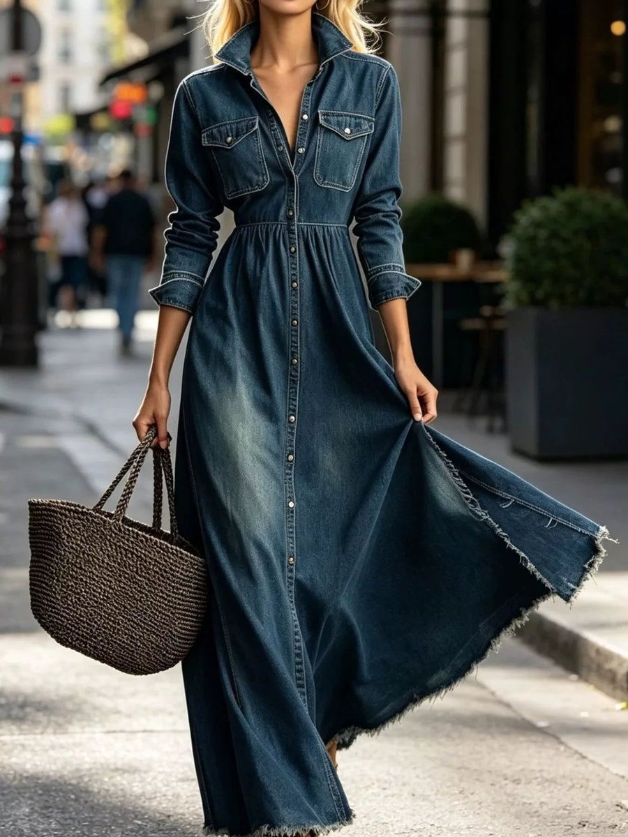 A woman wearing a dark blue denim maxi dress with a button-front closure and chest pockets, walking on a city street while carrying a woven tote bag.