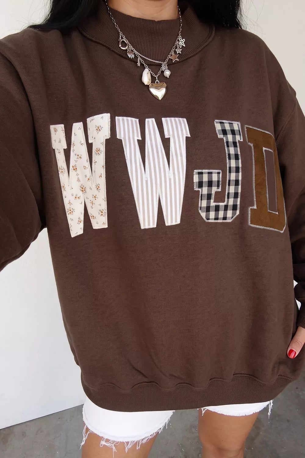 A person wearing a brown sweatshirt with large decorative letters 'WWJD' across the chest in mixed patterns, paired with white frayed shorts and layered necklaces.