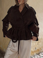Person wearing a brown peplum jacket with a belt, standing against a textured wall.