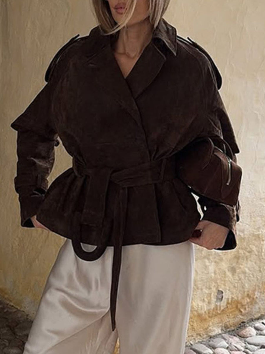 Person wearing a brown peplum jacket with a belt, standing against a textured wall.