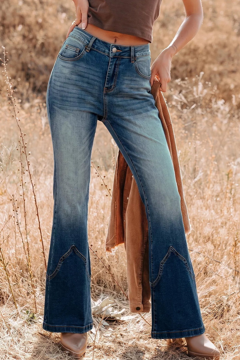 A person standing in a dry grassy field wearing blue denim flare jeans with a faded wash and decorative stitching near the lower legs. A brown top and tan boots are also visible, along with a brown jacket draped over one arm.