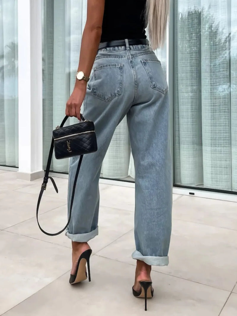 Person wearing light blue jeans and black high-heeled shoes, holding a black handbag indoors.
