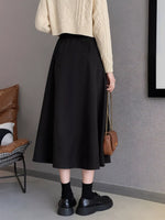 Person wearing a beige sweater, black skirt, and black shoes in a room with a brown chair and gray wall.