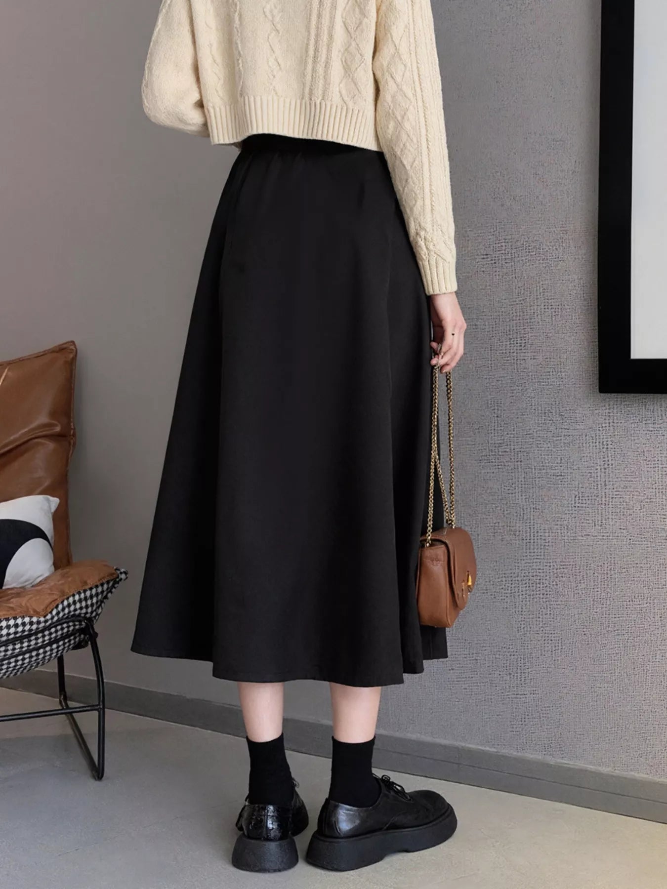 Person wearing a beige sweater, black skirt, and black shoes in a room with a brown chair and gray wall.