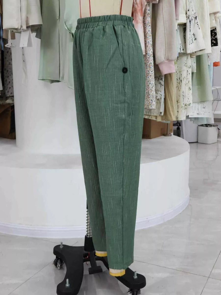 Green wide-leg trousers with an elastic waistband and a single black button on the left side, displayed on a dress form in a clothing store.