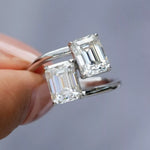 Silver ring with emerald-cut diamonds held by a hand against a gray background