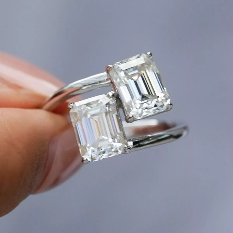 Silver ring with emerald-cut diamonds held by a hand against a gray background
