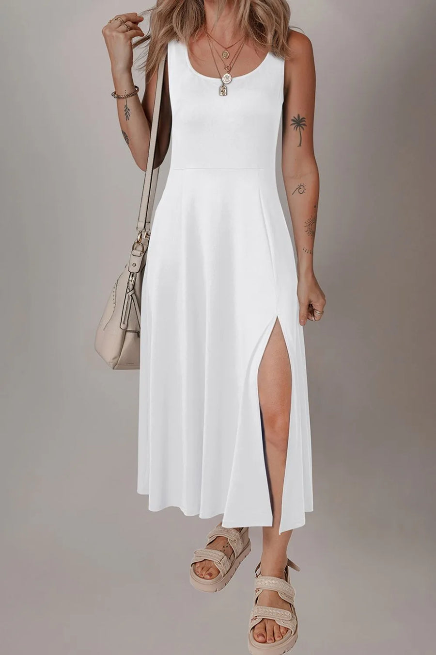 Woman wearing a white sleeveless dress with a slit, holding a beige handbag against a plain background.