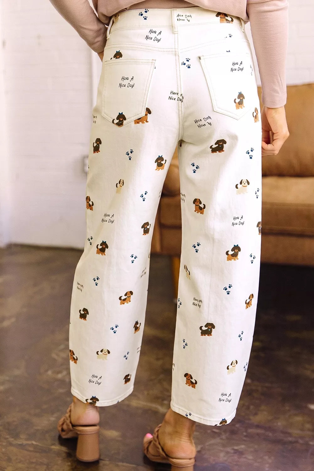 A person shown from the back wearing off-white wide-leg cropped pants with a repeating print of small brown dachshunds, blue paw prints, and handwritten-style text, paired with tan heeled sandals in an indoor room setting.